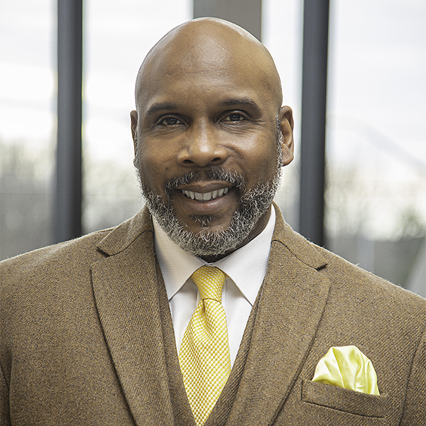 Photo of Omar Eaton-Martínez wearing a brown suit and a white shirt paired with a yellow tie with pocket square. He is bald, dark skinned, and salt and pepper bearded. He's smiling broadly and looking at the camera.